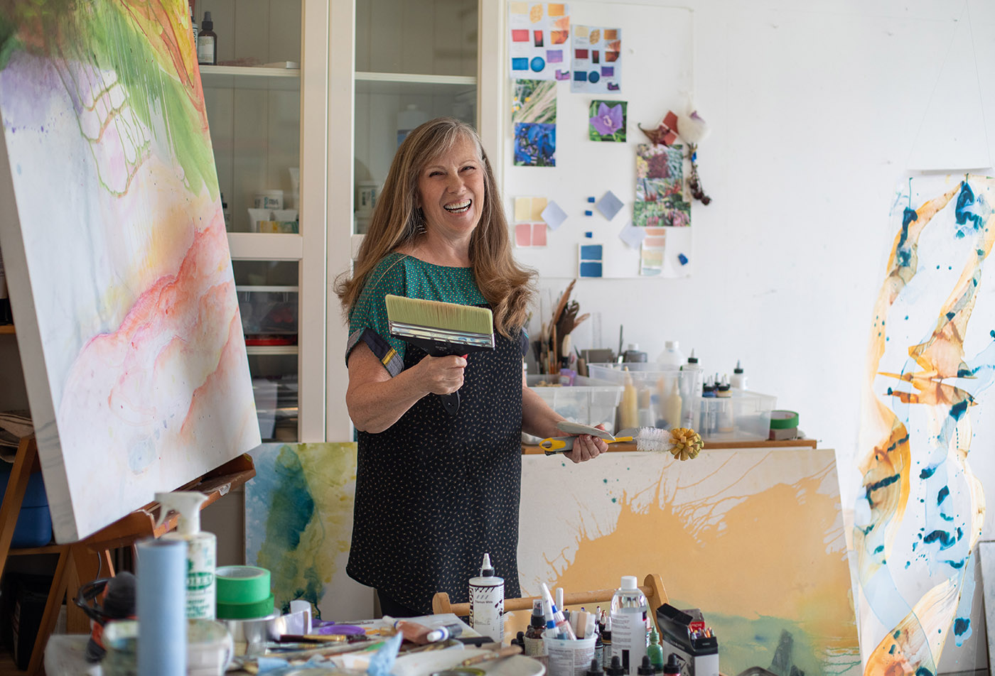Jan Jensen holding a large paint brush and smiling in her studio 